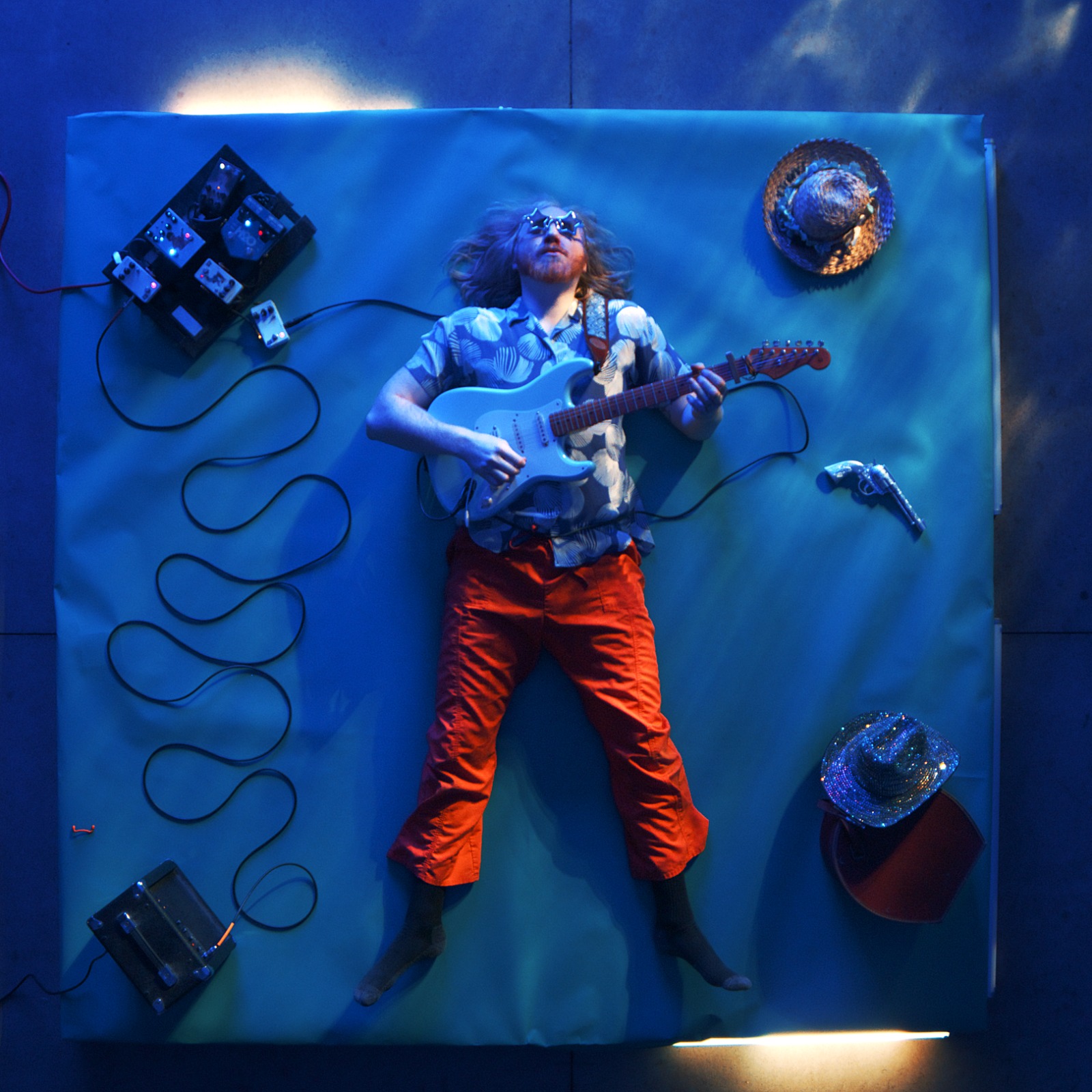 Dean Vision lying on a blue backdrop with a guitar, pedalboard, hats and orange pants — the Parallel Play album cover